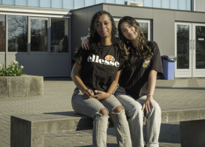 Twee jonge vrouwen zitten op een bank voor een schoolgebouw.