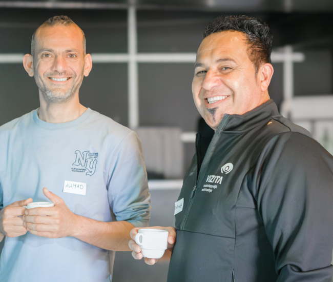 Twee mannen staan lachend met een kopje koffie in de hand.