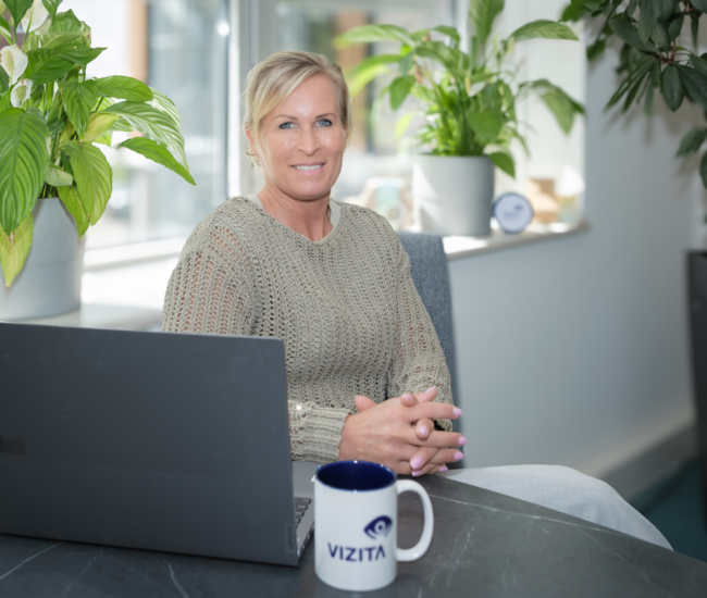 Vrouw zit aan bureau met laptop en koffie, omringd door planten.