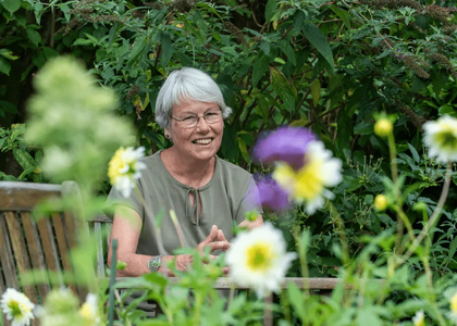 Een glimlachende vrouw zit tussen kleurrijke bloemen in een tuin.
