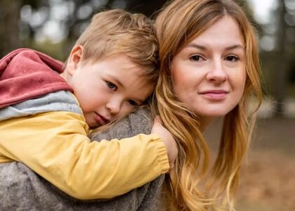 Een moeder draagt haar kind op de rug in een bosrijke omgeving.