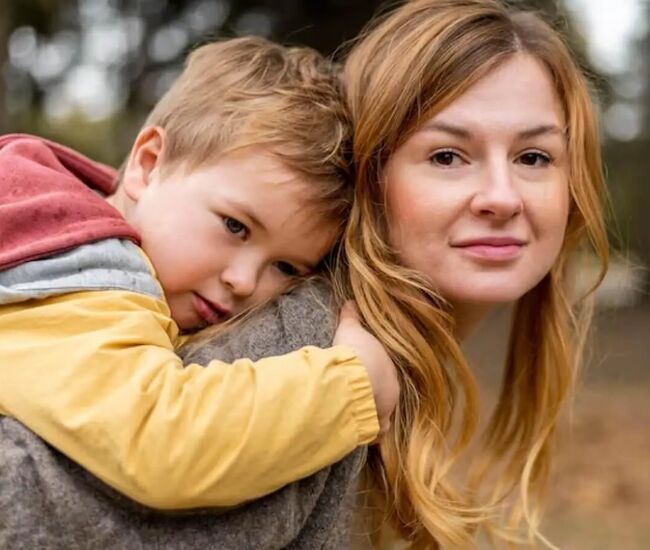 Een moeder draagt haar kind op de rug in een bosrijke omgeving.