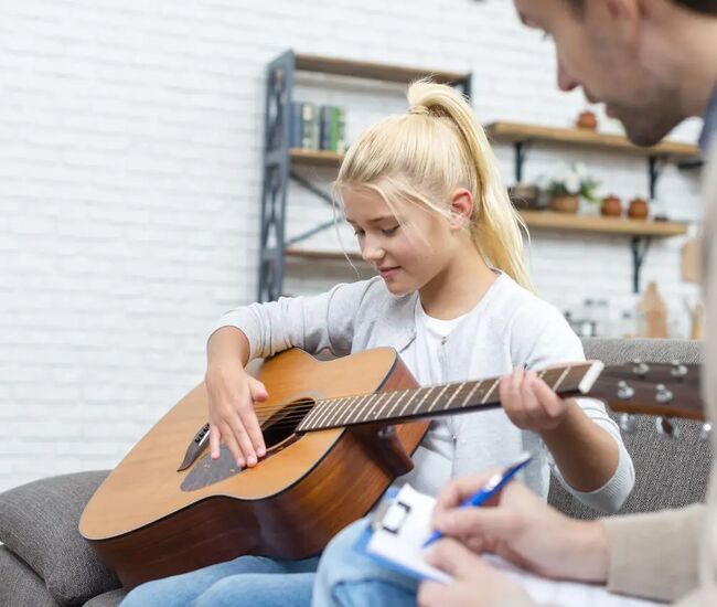 Een meisje speelt gitaar terwijl een volwassene aantekeningen maakt.