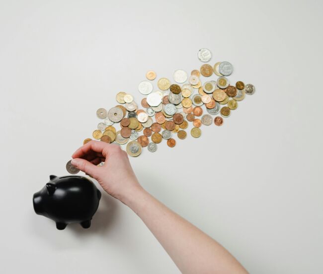Een hand plaatst een munt in een zwart spaarvarken, omringd door munten.