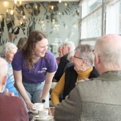 Een jongere serveert koffie aan een groep oudere mensen in een café.