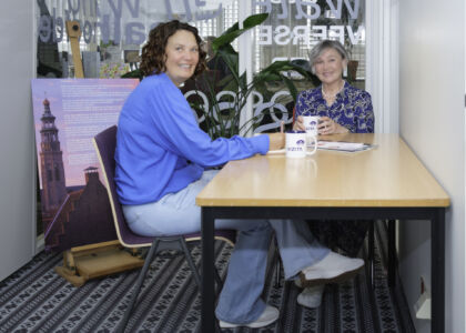 Twee vrouwen zitten aan een tafel met koffiemokken en praten.