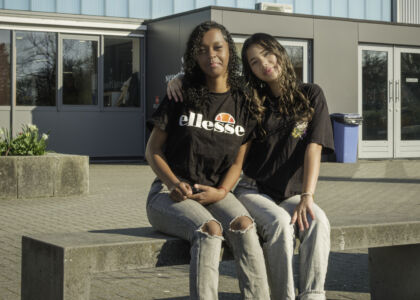 Twee jonge vrouwen zitten op een bank voor een schoolgebouw.