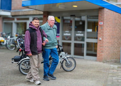Twee mannen lopen samen buiten een gebouw met fietsen op de achtergrond.