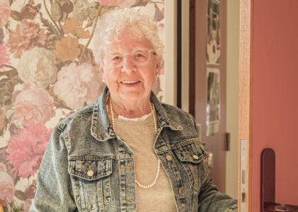 Oudere vrouw met krullend haar staat lachend bij een deur.