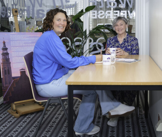 Twee vrouwen zitten aan een tafel met koffiemokken en praten.