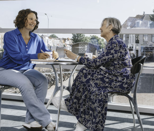 Twee vrouwen zitten aan een tafel en hebben een gesprek met koffie.