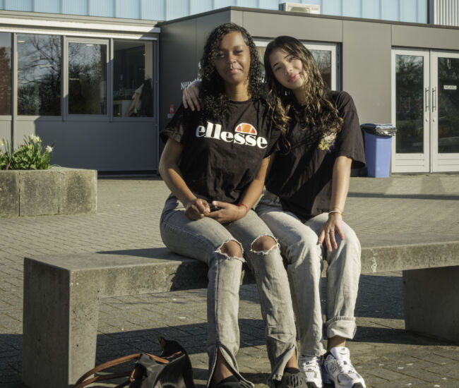 Twee jonge vrouwen zitten op een bank voor een schoolgebouw.