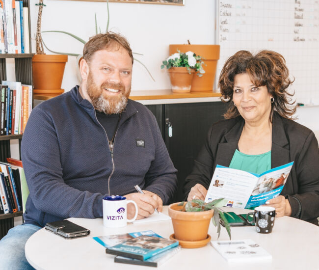 Twee mensen zitten aan een tafel met boeken en een plant.