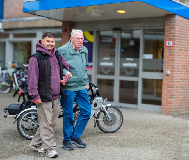 Twee mannen lopen samen buiten een gebouw met fietsen op de achtergrond.