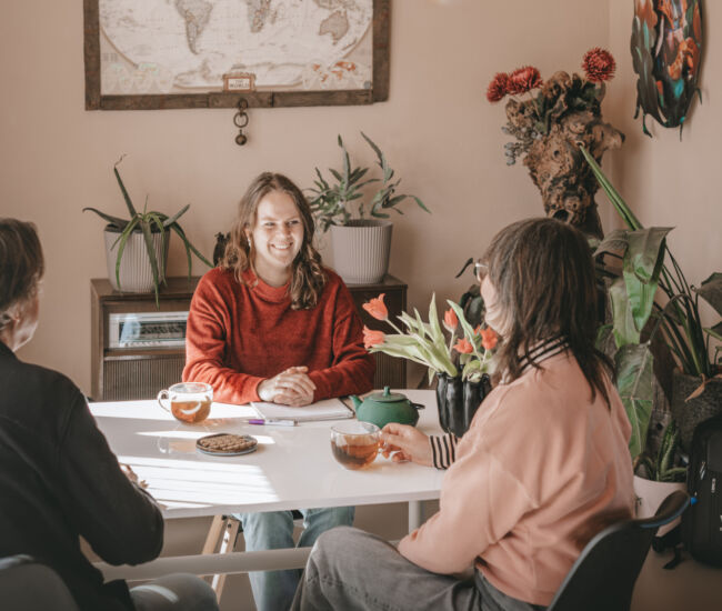 Drie mensen zitten aan tafel met thee en bloemen.