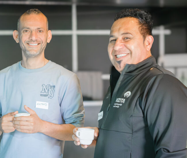 Twee mannen staan lachend met een kopje koffie in de hand.