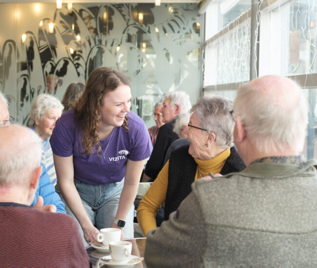 Een jongere serveert koffie aan een groep oudere mensen in een café.