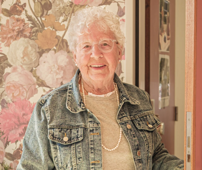 Oudere vrouw met krullend haar staat lachend bij een deur.