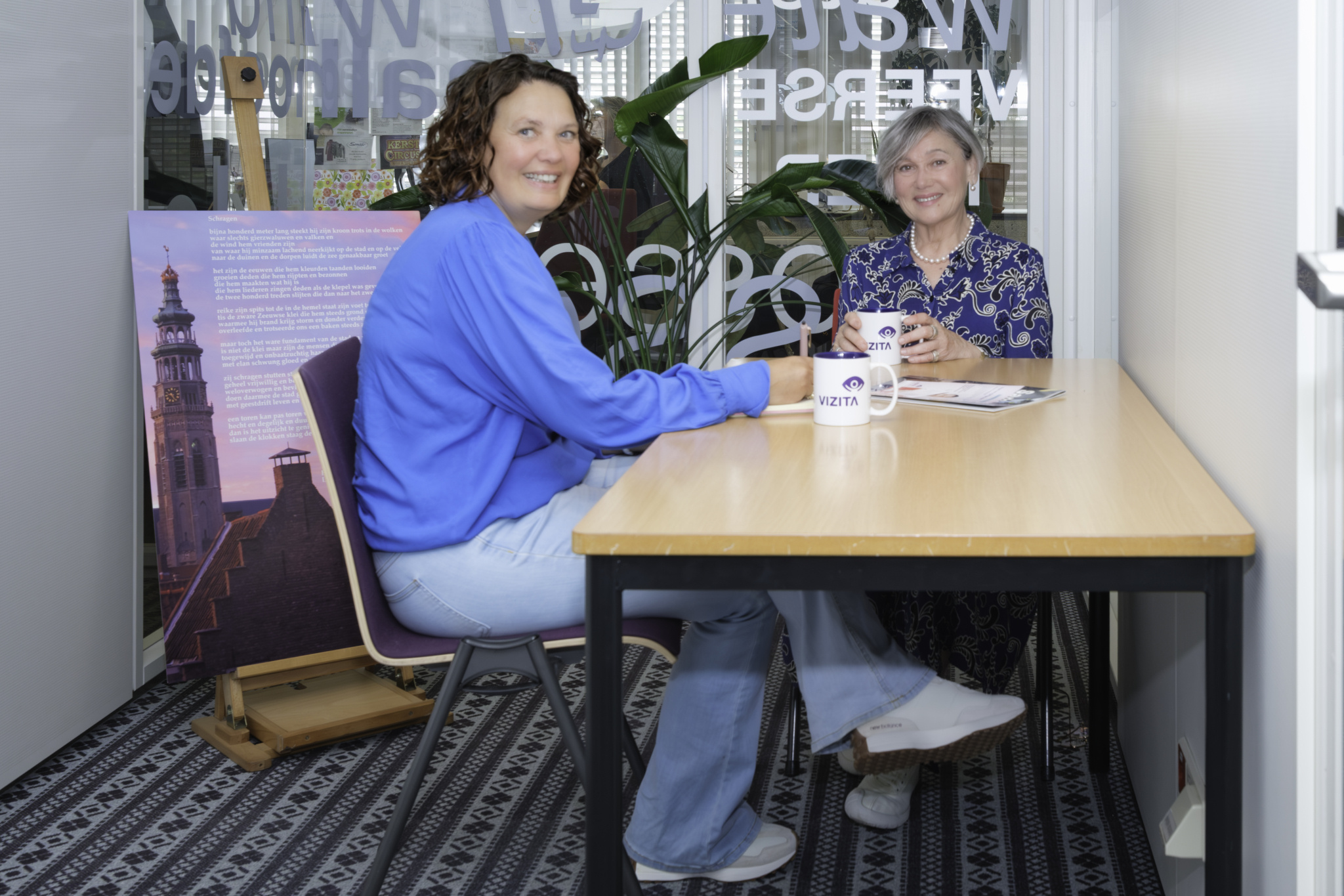 Twee vrouwen zitten aan een tafel met koffiemokken en praten.