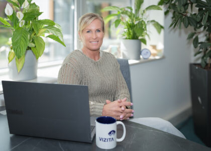 Vrouw zit aan bureau met laptop en koffie, omringd door planten.
