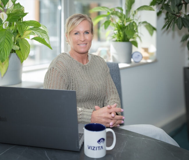 Vrouw zit aan bureau met laptop en koffie, omringd door planten.