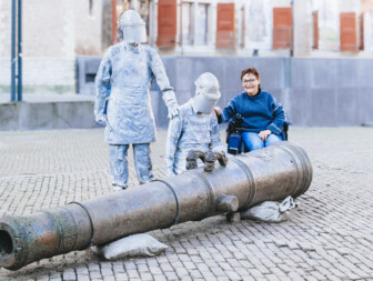 Vrouw in rolstoel met twee standbeelden bij een kanon.