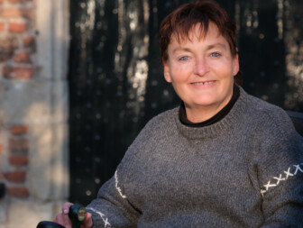 Vrouw in rolstoel met een grijze trui, glimlachend naar de camera.
