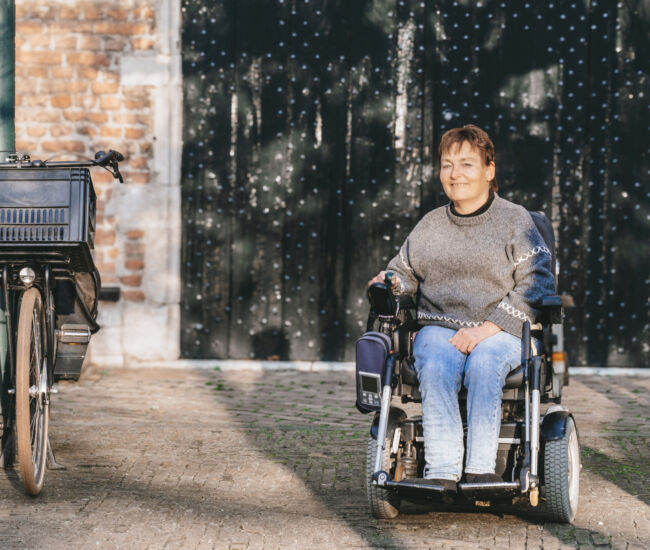 Vrouw in rolstoel poseert naast een fiets in een straat.