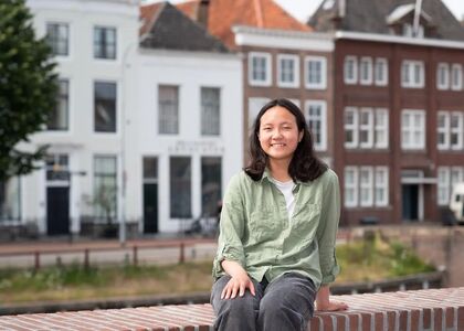 Jonge vrouw zit op een muurtje met historische gebouwen op de achtergrond.