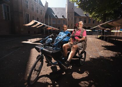 Twee mannen zitten op een tandemfiets in een zonnige straat.