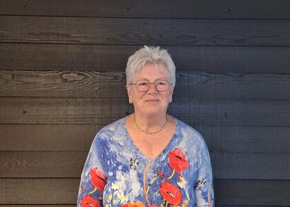 Vrouw met kort grijs haar en een kleurrijke blouse, staand voor een houten muur.