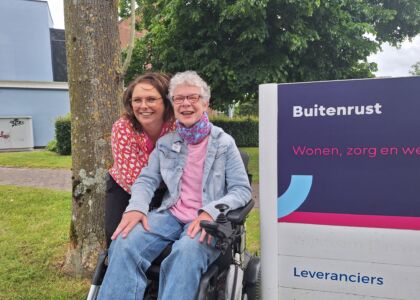 Twee vrouwen poseren lachend voor een zorginstelling met een bord.