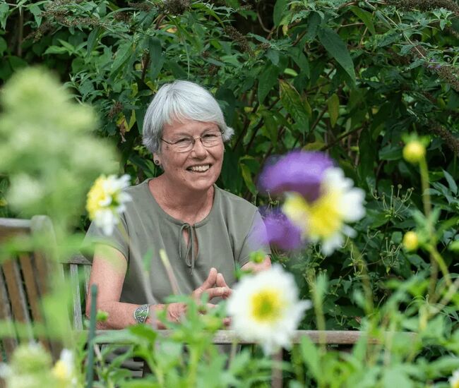 Een glimlachende vrouw zit tussen kleurrijke bloemen in een tuin.