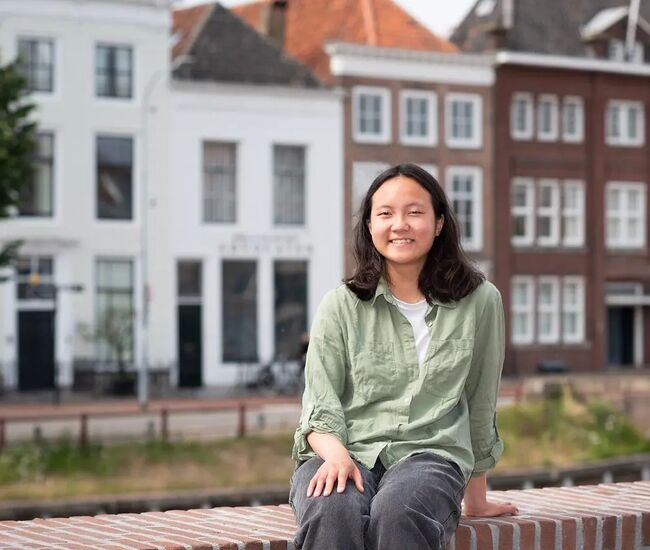 Jonge vrouw zit op een muurtje met historische gebouwen op de achtergrond.