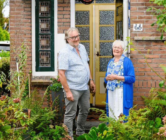 Twee mensen staan lachend voor een huis met een groene tuin.