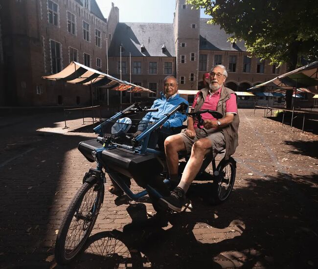 Twee mannen zitten op een tandemfiets in een zonnige straat.