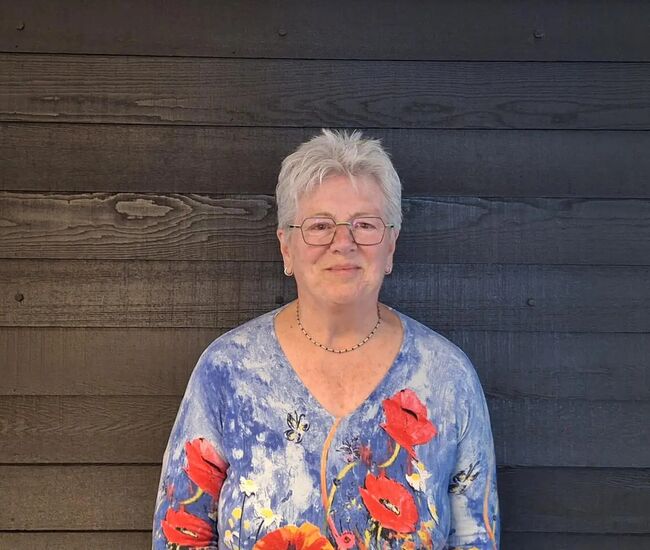 Vrouw met kort grijs haar en een kleurrijke blouse, staand voor een houten muur.