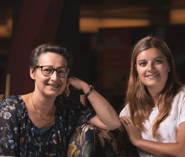Twee vrouwen zitten samen en lachen naar de camera.