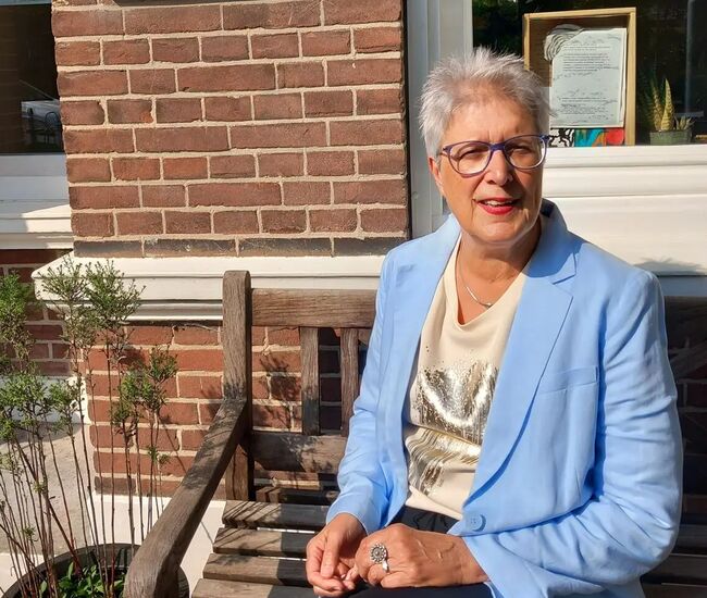 Vrouw in een lichtblauwe blazer zit op een houten bank.