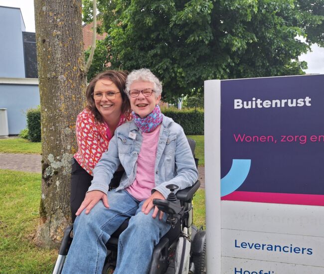 Twee vrouwen poseren lachend voor een zorginstelling met een bord.