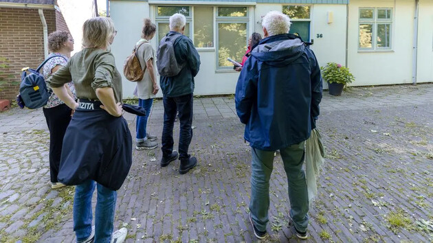 Groep mensen staat voor een huis en luistert naar uitleg.