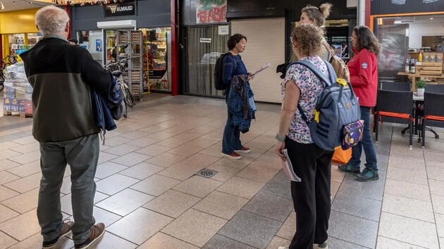 Groep mensen in een winkelcentrum, met een vrouw die iets uitlegt.