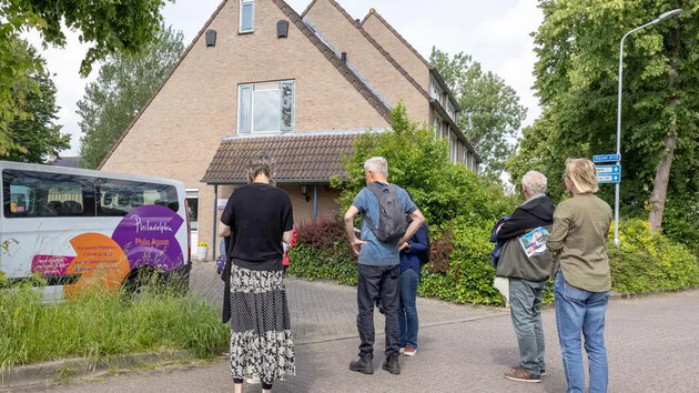 Groep mensen staat voor een huis met een busje ernaast.